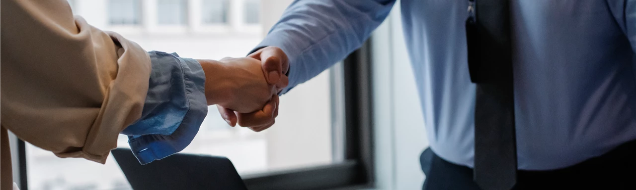 Crop unrecognizable coworkers in formal wear standing at table with laptop and documents while greeting each other before meeting