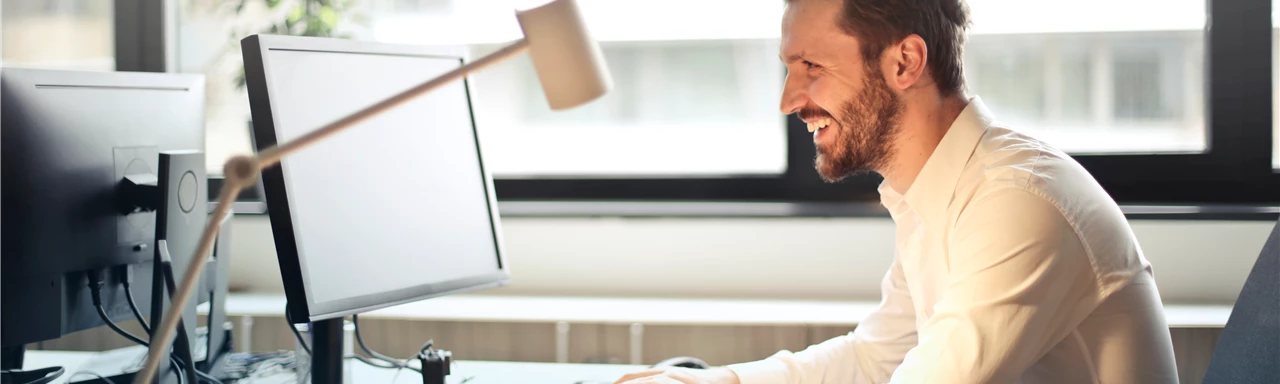 Man in White Dress Shirt Sitting on Black Rolling Chair While Facing Black Computer Set and Smiling